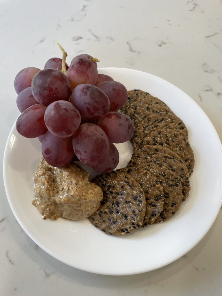 a white plate with black sesame crackers, almond butter and red grapes